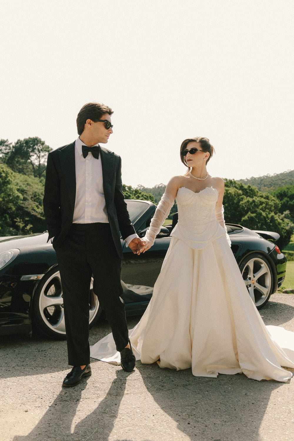 Groom in a black tuxedo and the bride by a Porsche